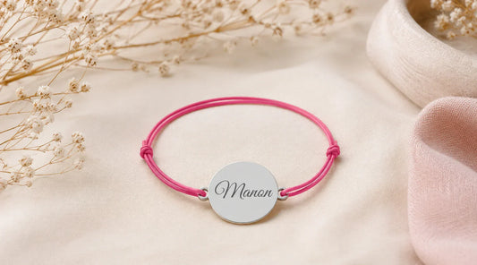 Bracelet cordon personnalisé avec médaille en argent gravée d’un prénom, présenté sur un tissu beige dans une ambiance élégante.