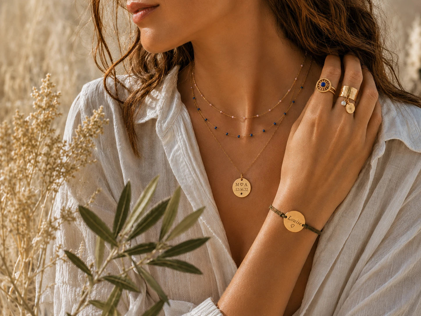 Femme portant plusieurs colliers dorés avec perles bleues et roses, une médaille gravée, des bagues et un bracelet cordon personnalisé avec le prénom Louise.