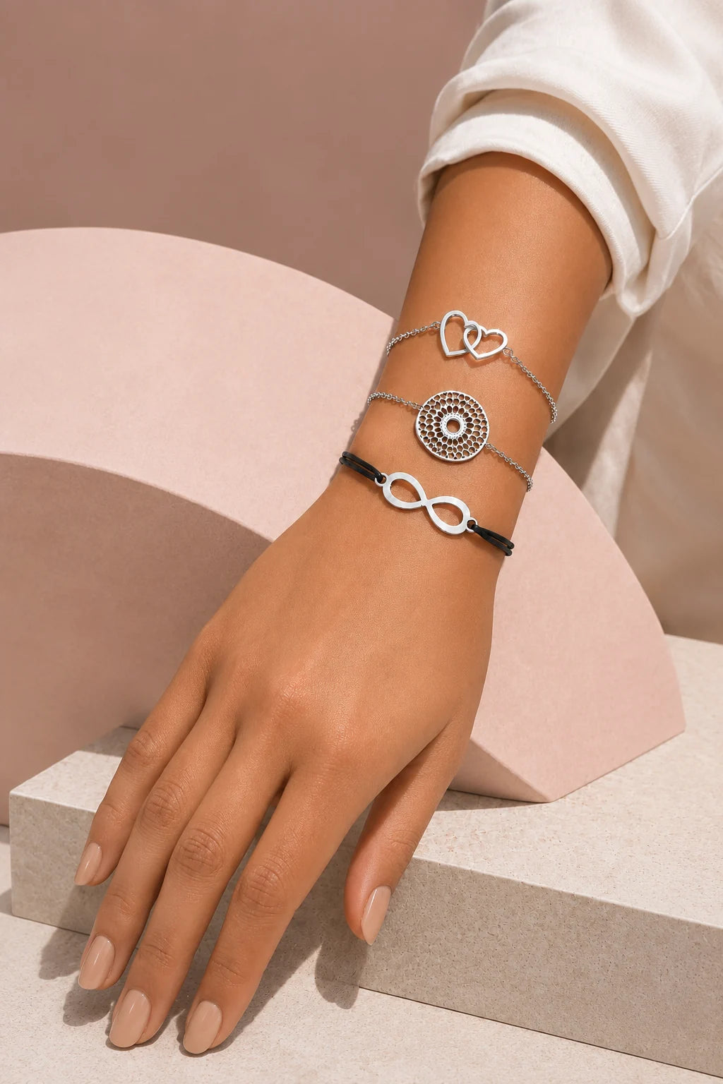 Main portant trois bracelets argentés, dont un bracelet cœurs, un bracelet rond ajouré et un bracelet infini sur fond beige rosé.